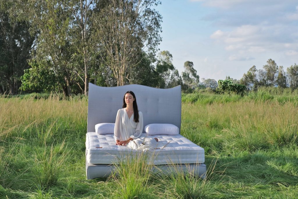 a female model ready to sleep in a luxury bed and mattress with two pillows in a green field with trees in the background showing sustainability