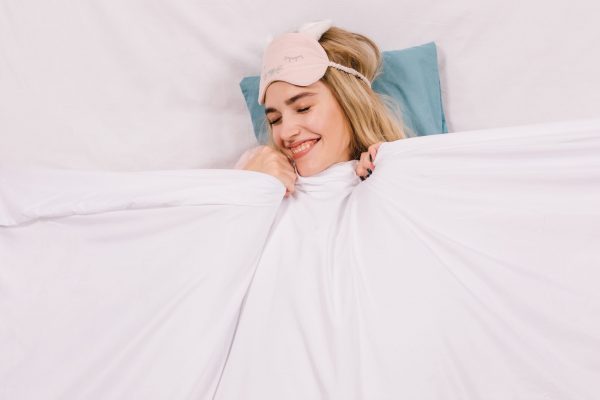 Carefree smiling girl lying in bed. Overhead shot of positive blonde woman in sleep mask.